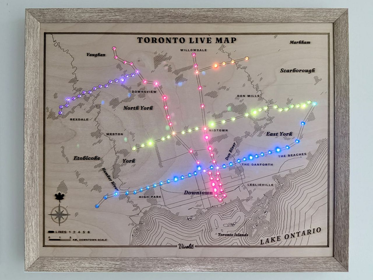 The Toronto Live Map hanging on a wall in a light wooden frame: an engraved birch map of Toronto with coloured lights along every subway line, neighbourhood names, and Lake Ontario drawn in contour lines